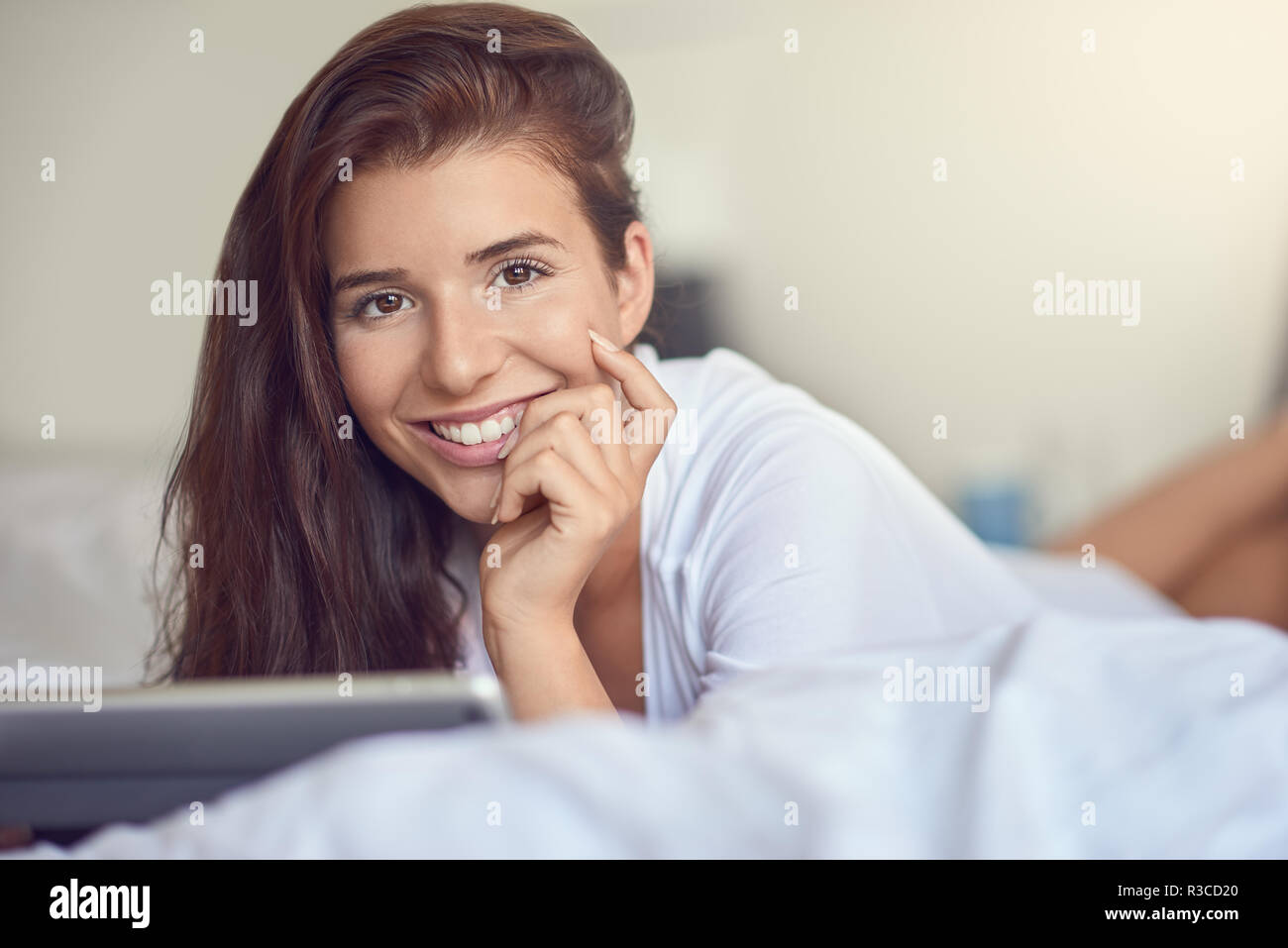 Pretty young woman with long brown hair, laying on stomach in bed with white linens and using tablet pc, smiling at the camera with copy space Stock Photo
