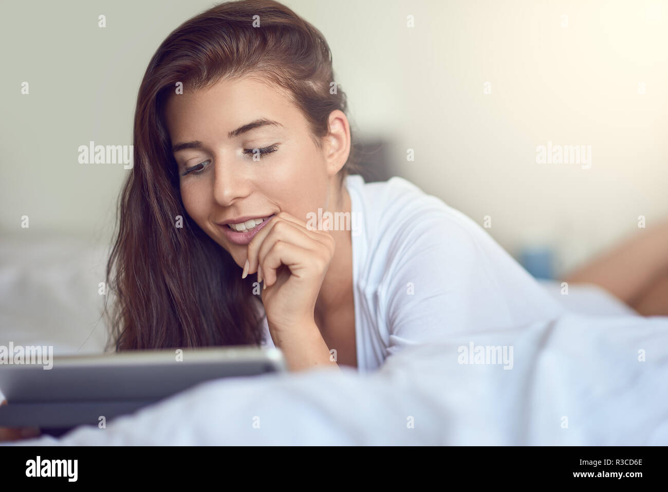 Pretty young woman with long brown hair, laying on stomach in bed with white linens and using tablet pc, doing home shopping or reading an e-mail Stock Photo