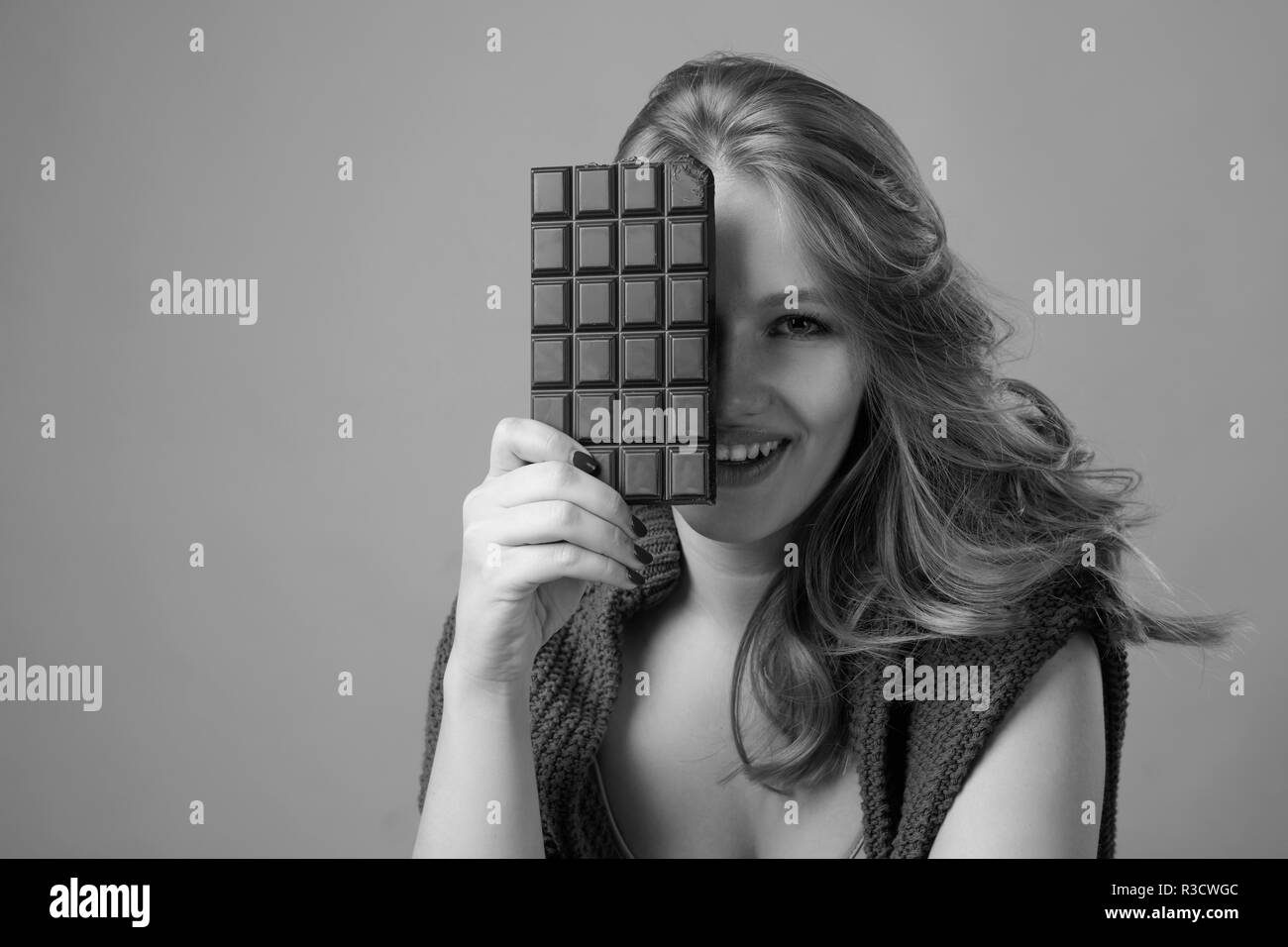 Happy young beautiful woman eating chocolate. Young blonde with natural makeup having fun and eating chocolate isolated on gray background. Stock Photo