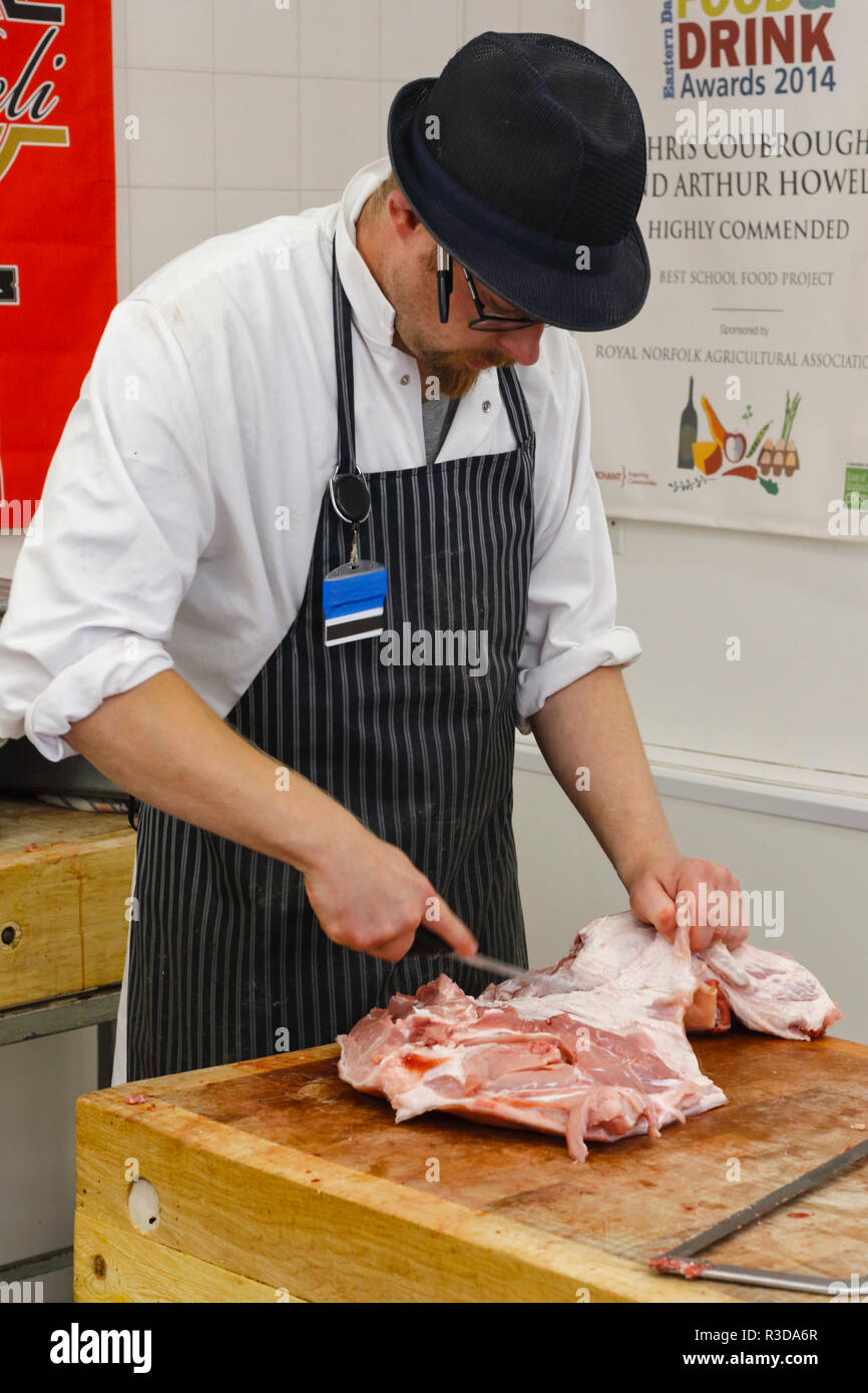 Butcher wearing a hat, vertical stripped apron, and a white coat preparing a joint of raw meat using a sharp butchers knife. England, UK Stock Photo