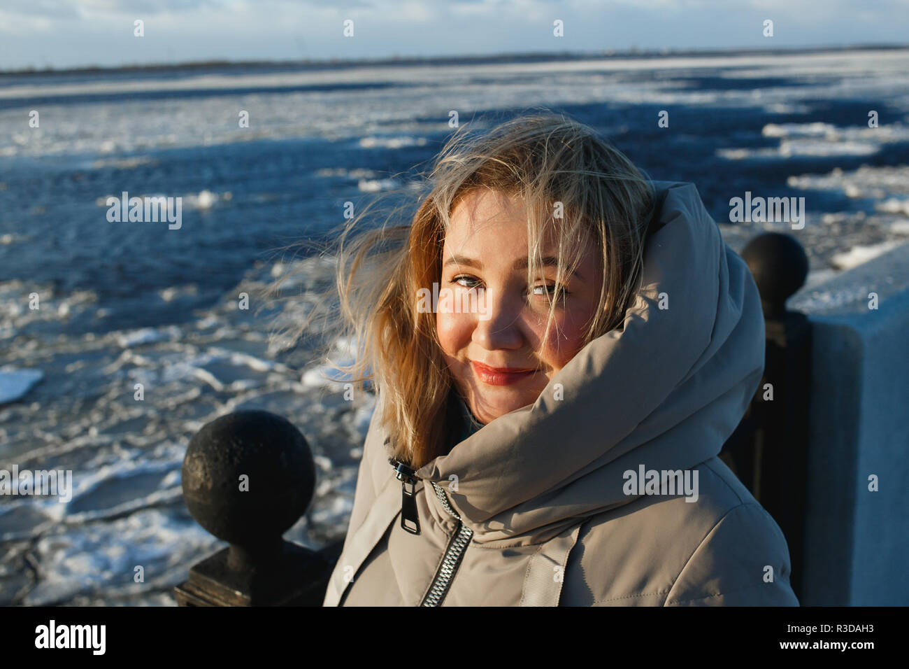 Portrait of pretty cheerful smiling woman on frozen river background in sunny winter day. Healthy winter walking in sinny frosty day. Stock Photo
