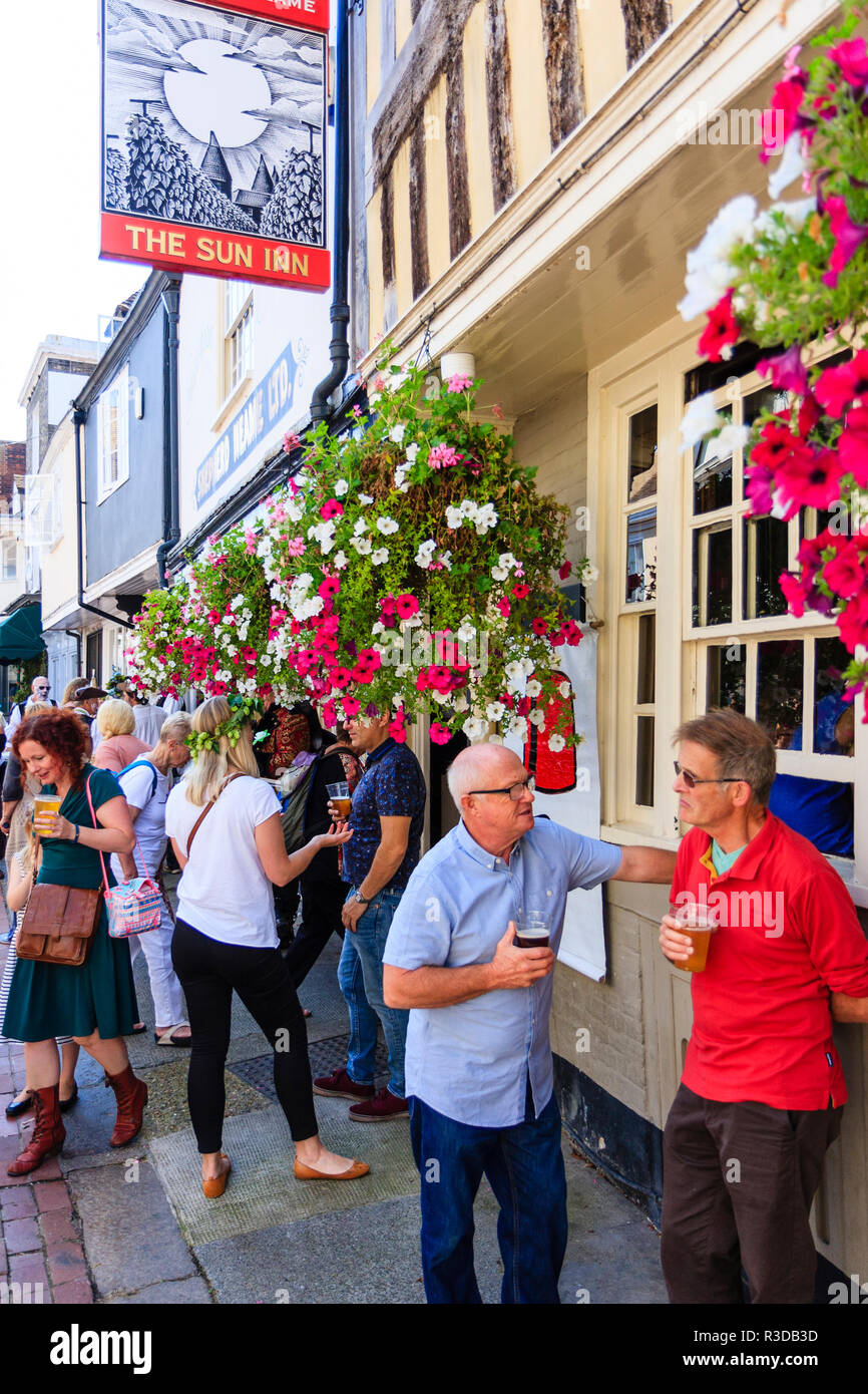 Two senior men standing chatting and holding beer glasses with other people behind, outside English town public house, with flowers hanging down. Stock Photo