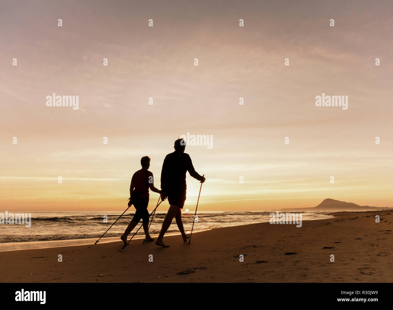 Mature couple walking with poles, Nordic walking, on beach at sunrise in Spain. Stock Photo