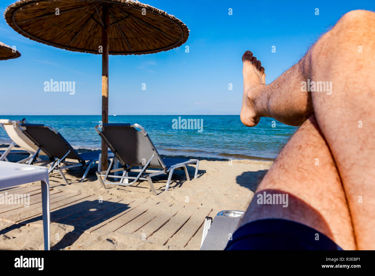 Man's legs until is sunbathing by lying carefree in lounger next to the coastline, on public beach. Stock Photo