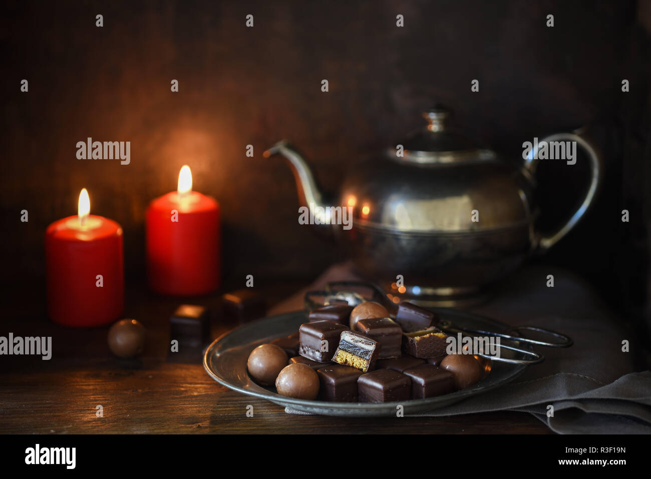 christmas chocolate sweets, two red candles and a silver teapott on a rustic wooden table, dark moody background, selected focus, narrow depth of fiel Stock Photo