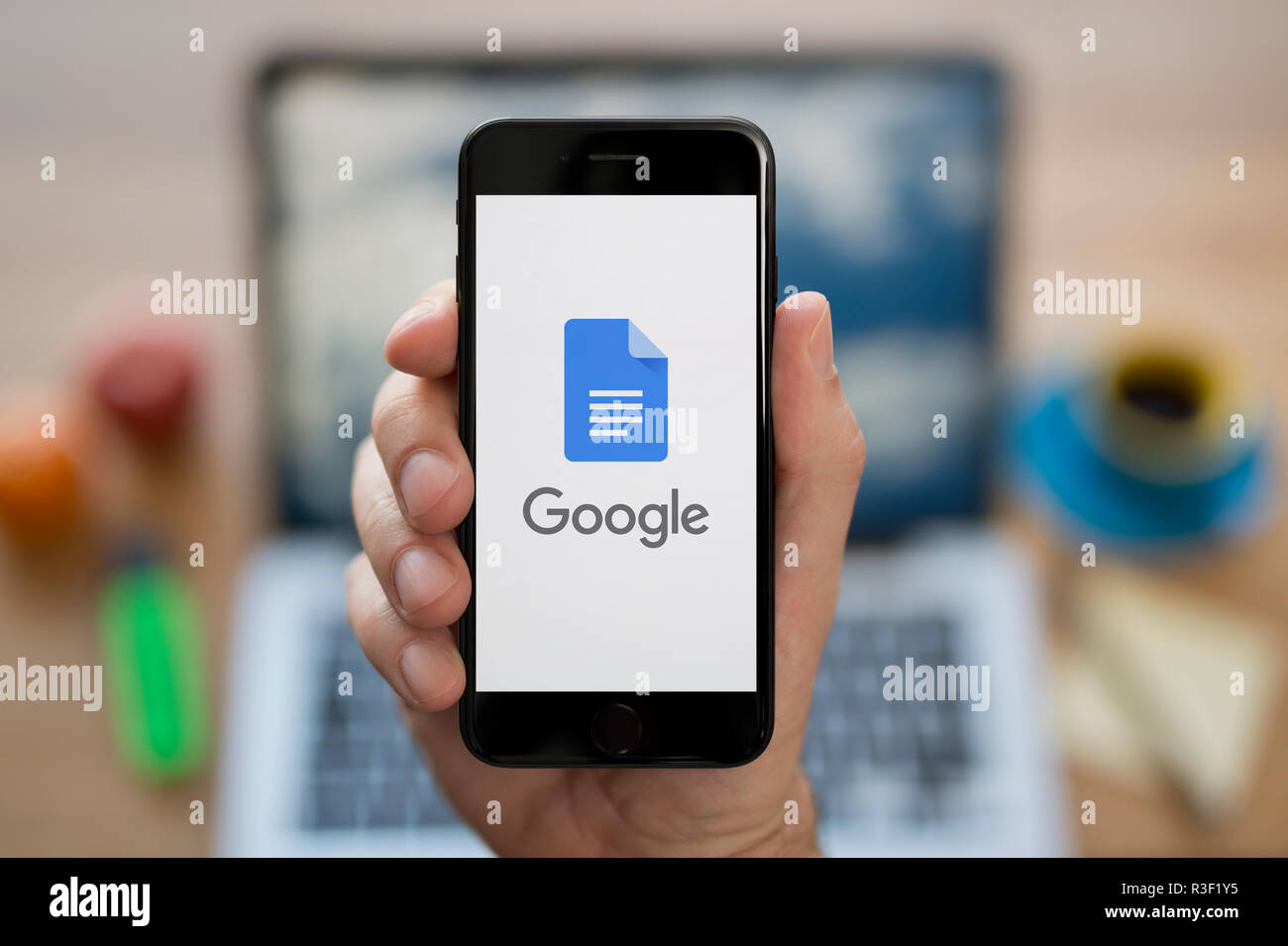 A man looks at his iPhone which displays the Google Docs logo, while sat at his computer desk (Editorial use only). Stock Photo