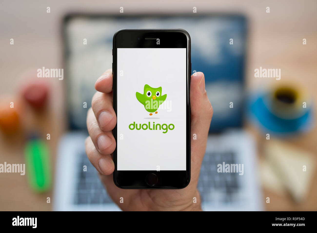 A man looks at his iPhone which displays the Duolingo logo, while sat at his computer desk (Editorial use only). Stock Photo