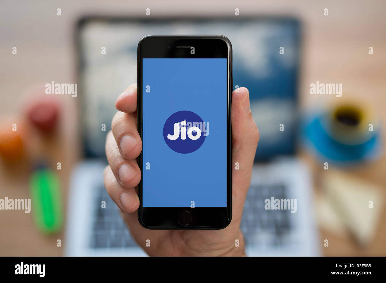A man looks at his iPhone which displays the Reliance Jio logo, while sat at his computer desk (Editorial use only). Stock Photo