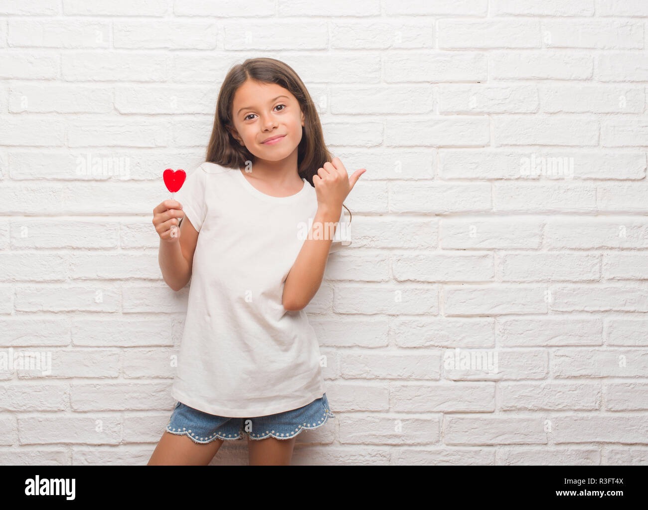 Young hispanic kid over white brick wall eating red heart lollipop