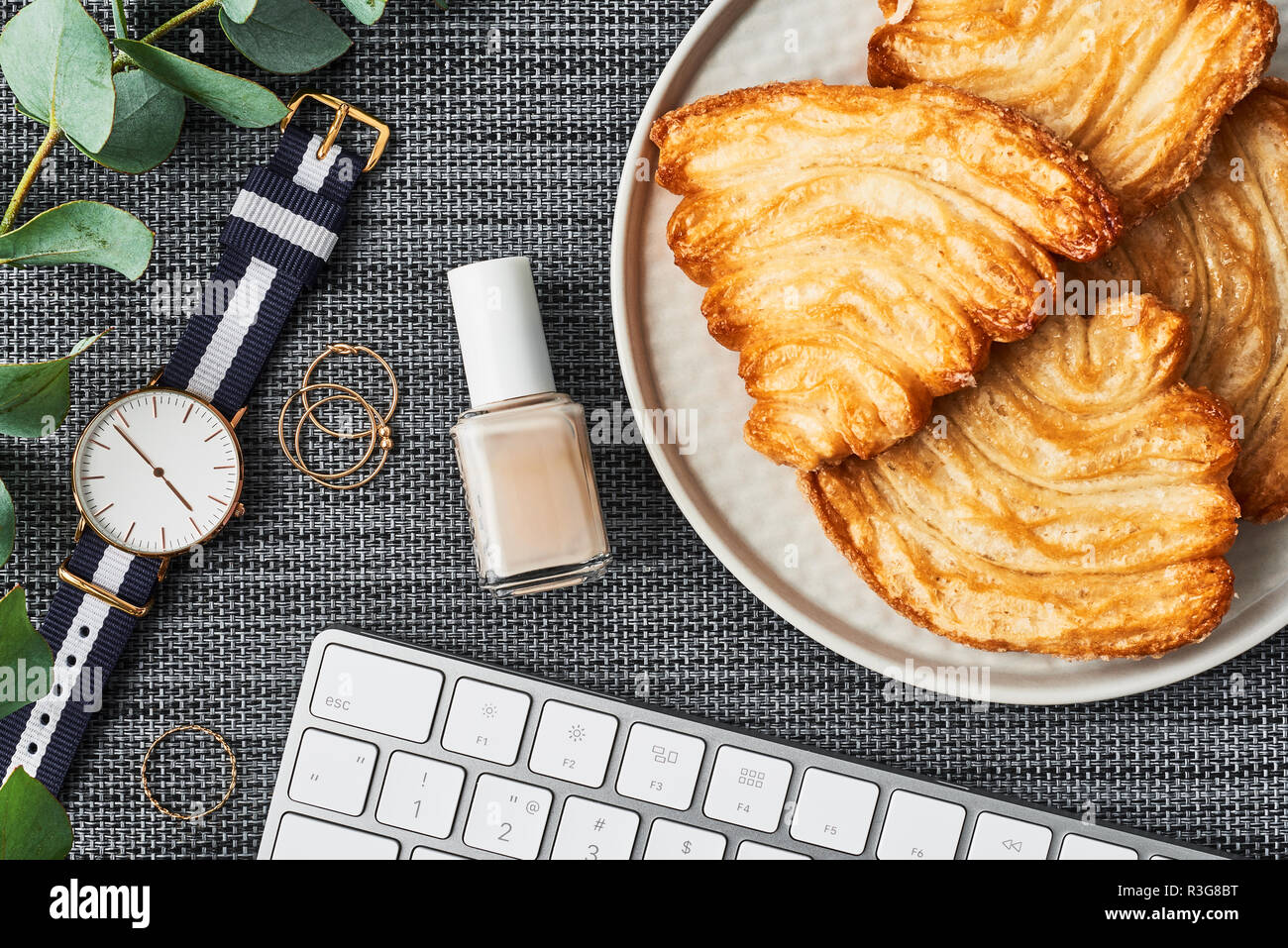 Cozy business composition with white computer keyboard, nail polish, eucalyptus plant, women's watch and large biscuits on grey background. Lunch brea Stock Photo