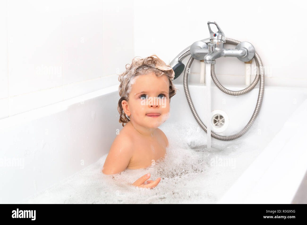A little girl is bathing in the bathroom with foam. Adorable baby girl with soap suds on hair taking bath. Closeup portrait of smiling kid, health car Stock Photo