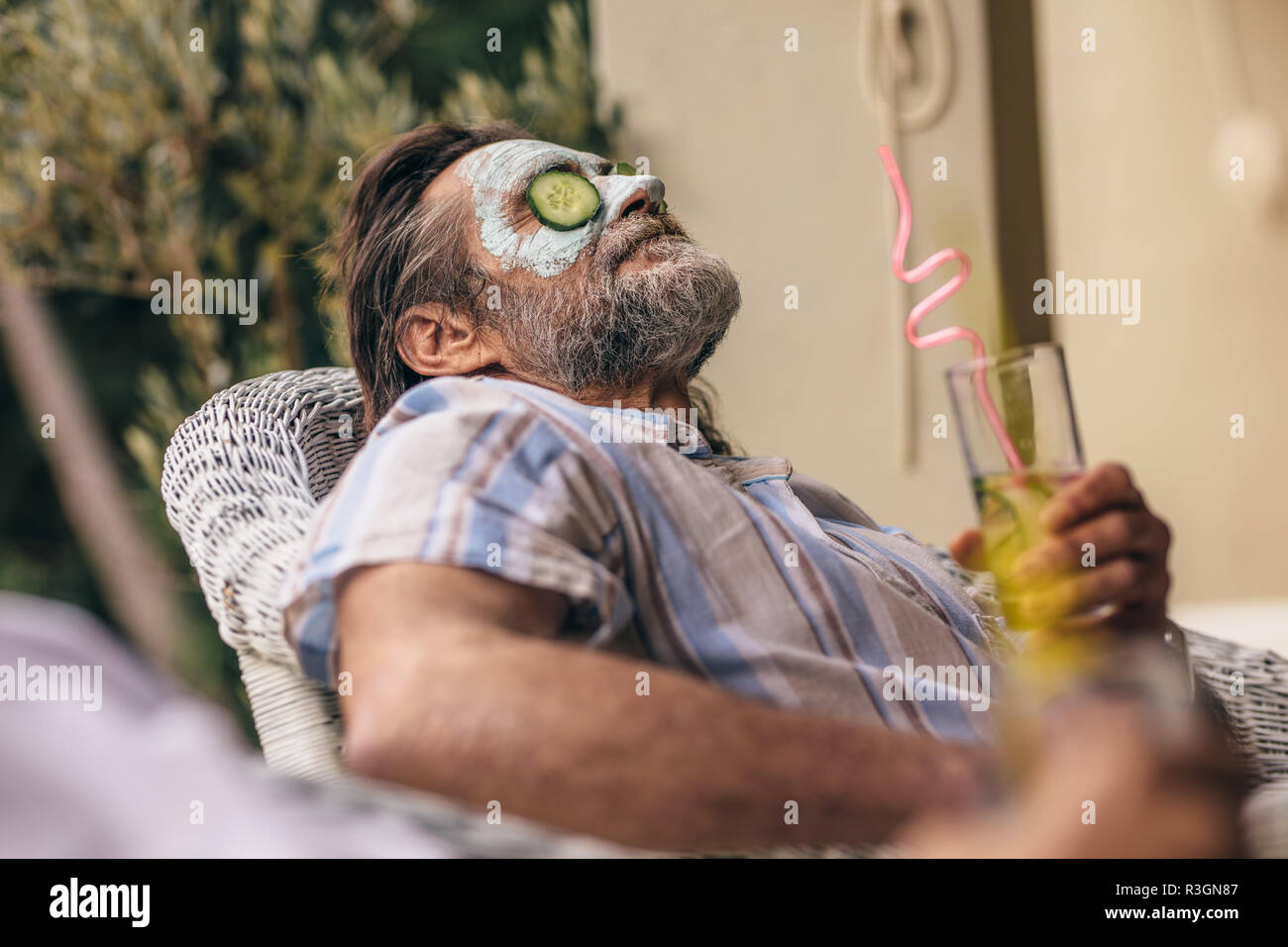 Relaxed elderly man sitting on chair with facial mask on face holding a glass of juice. Retired man relaxing with clay mask on face. Stock Photo