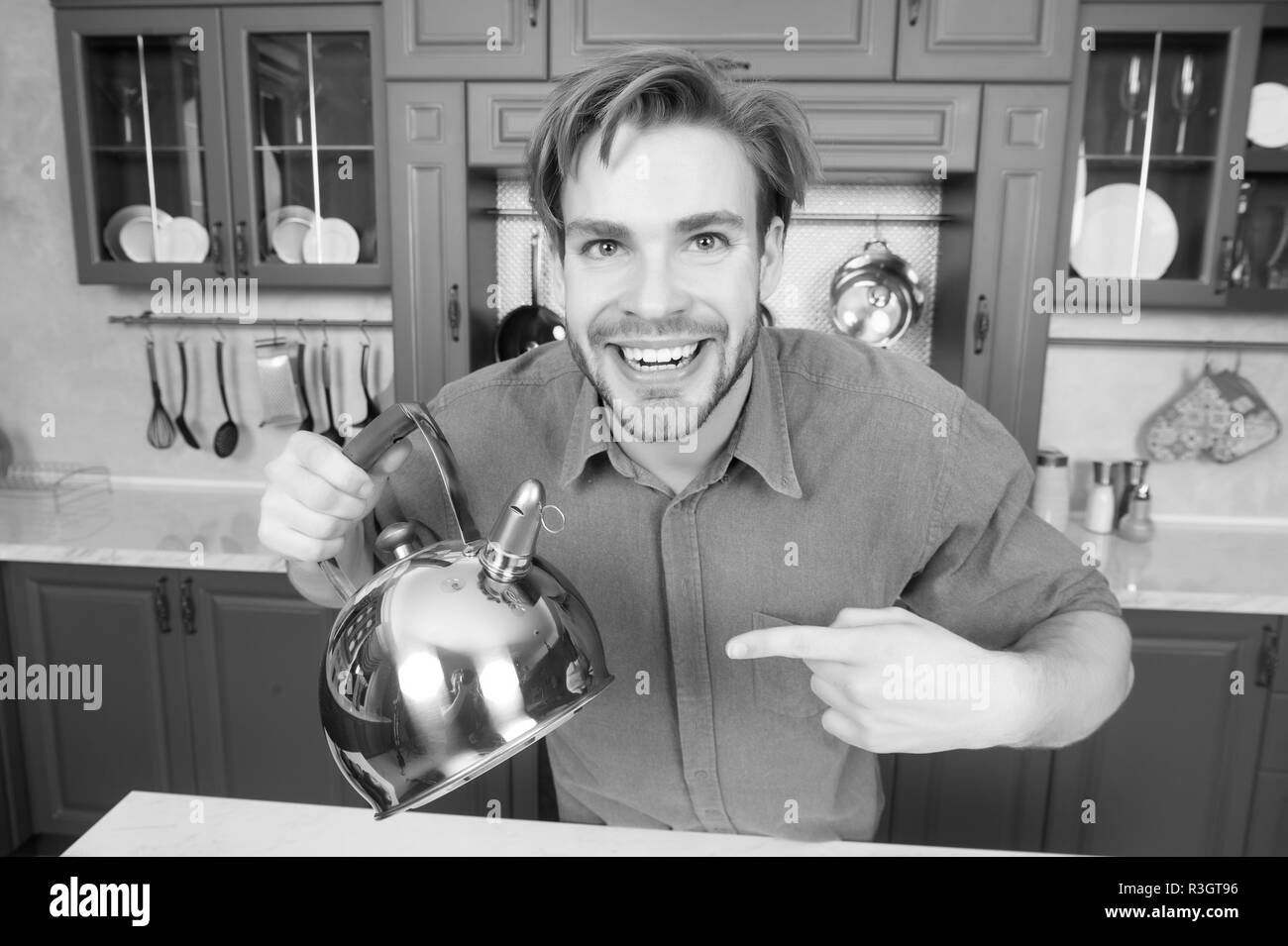 Happy man in blue shirt point finger at kettle. Kitchen appliance and furniture. Food preparation, cooking, heating, boiling water. Tea time, tea mood Stock Photo