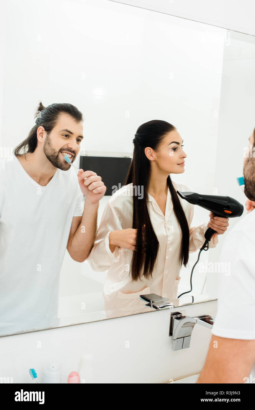 handsome adult man brushing teeth while wife drying hair with hairdryer Stock Photo