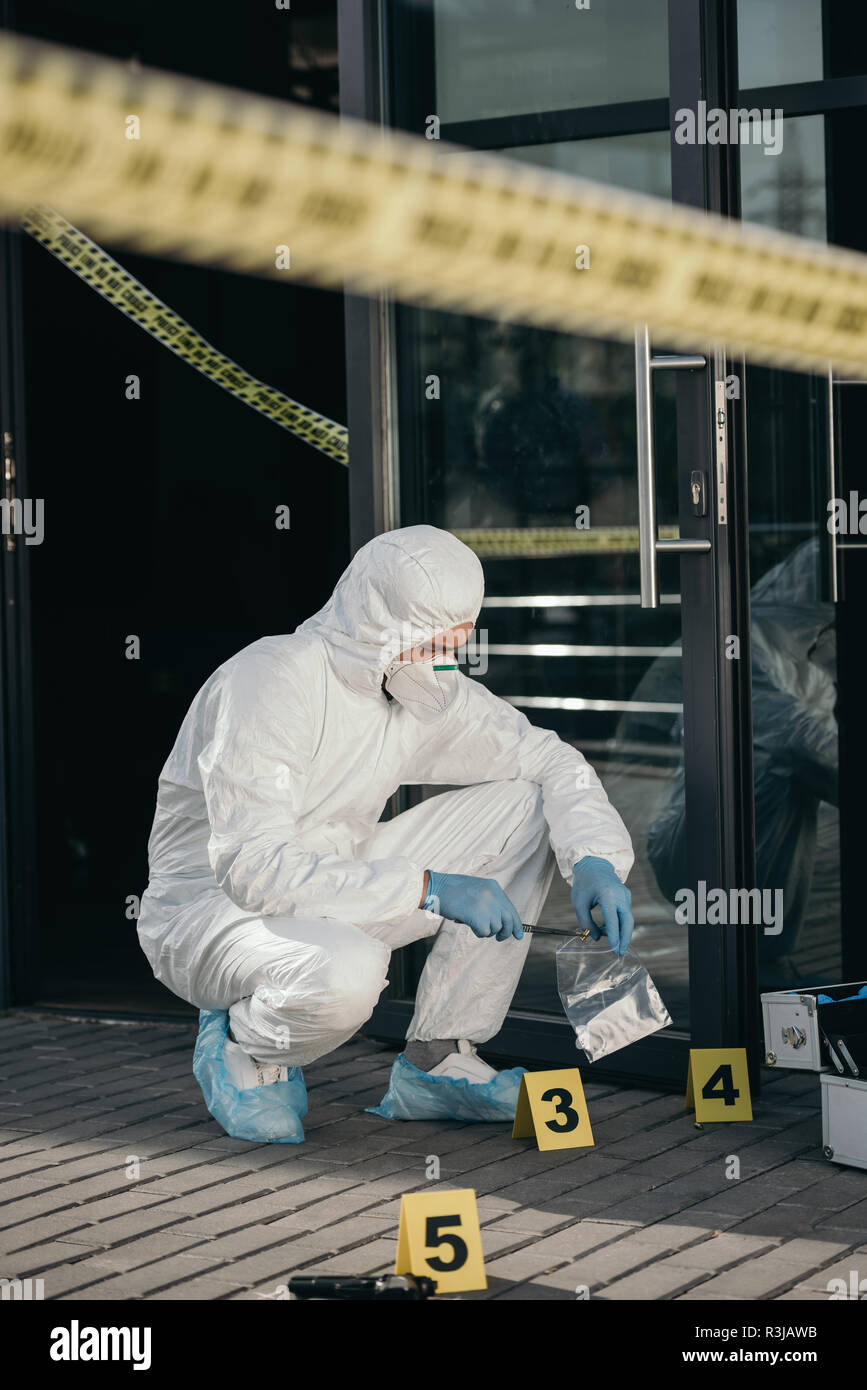 male criminologist in protective suit and latex gloves packing evidence with wizzles at crime scene Stock Photo