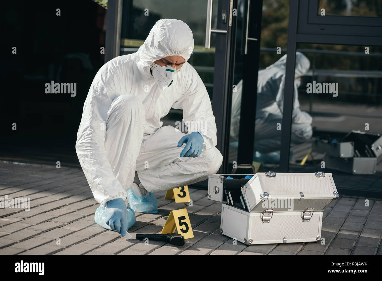 male criminologist in protective suit and latex gloves sitting near evidence gun Stock Photo