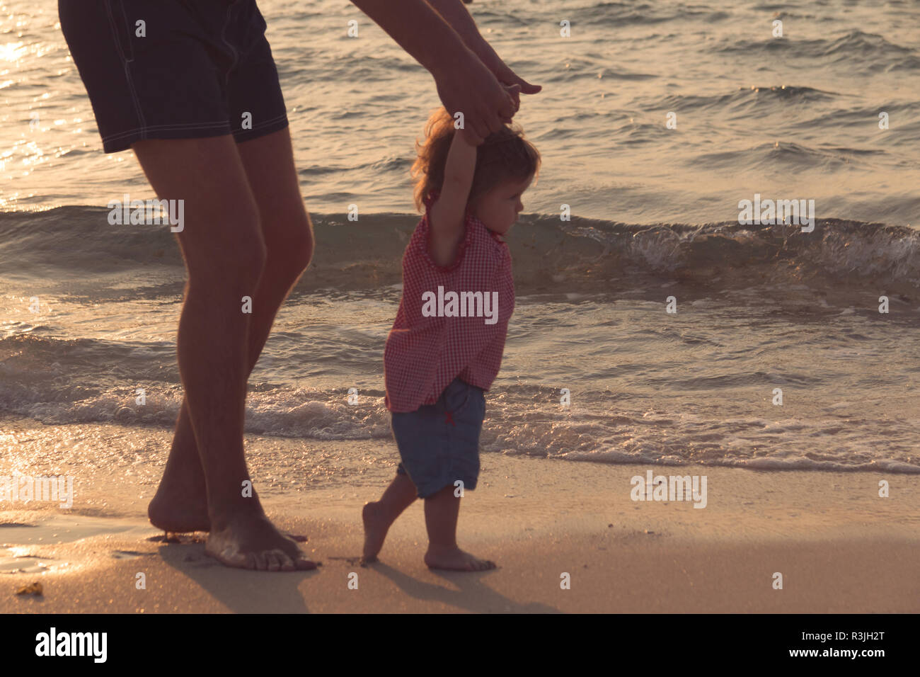 Father helping his baby with the first steps, teaching baby to walk concept, outdoor candid photo on the beach, spending a day at the beach, healthy family lifestyle. Stock Photo