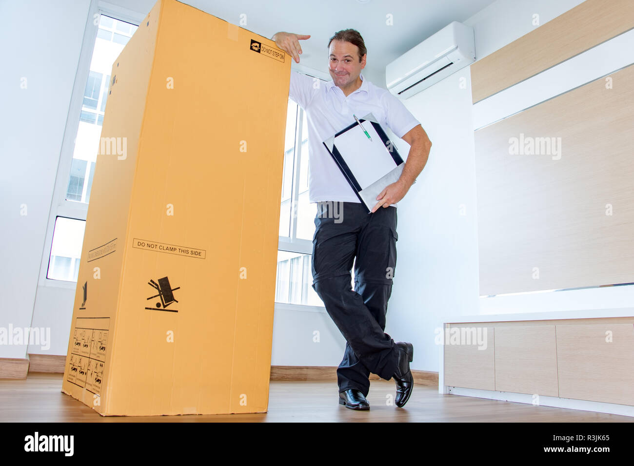 A man shows a large package in an empty room. The postman delivers the parcel to the new apartment. Stock Photo