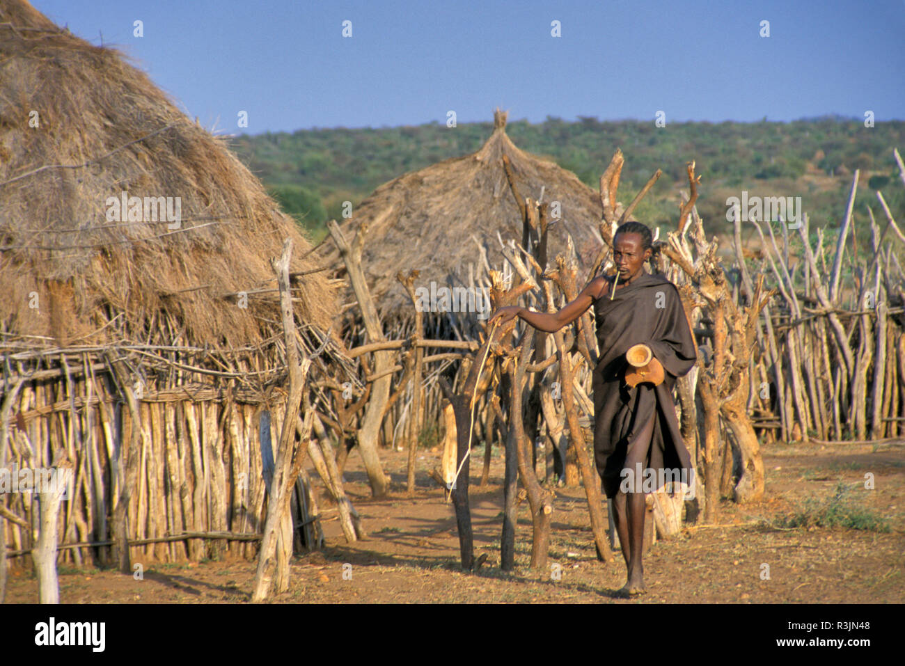 Africa, Ethiopia, Omo region. A Hamar tribe man with his portable wooden stool walks along a stick fence and thatched-roof village huts. (MR) Stock Photo