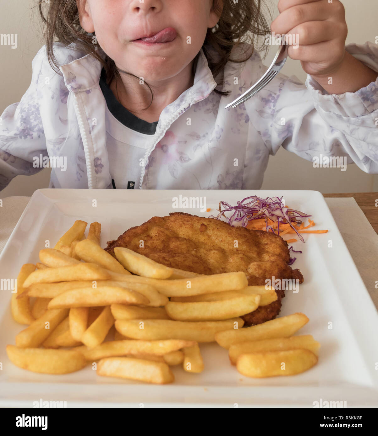Little italian girl eating breaded meat and French fries with a fork,  in a restaurant. The child is wearing flower jacket. Ideal for concepts. Stock Photo