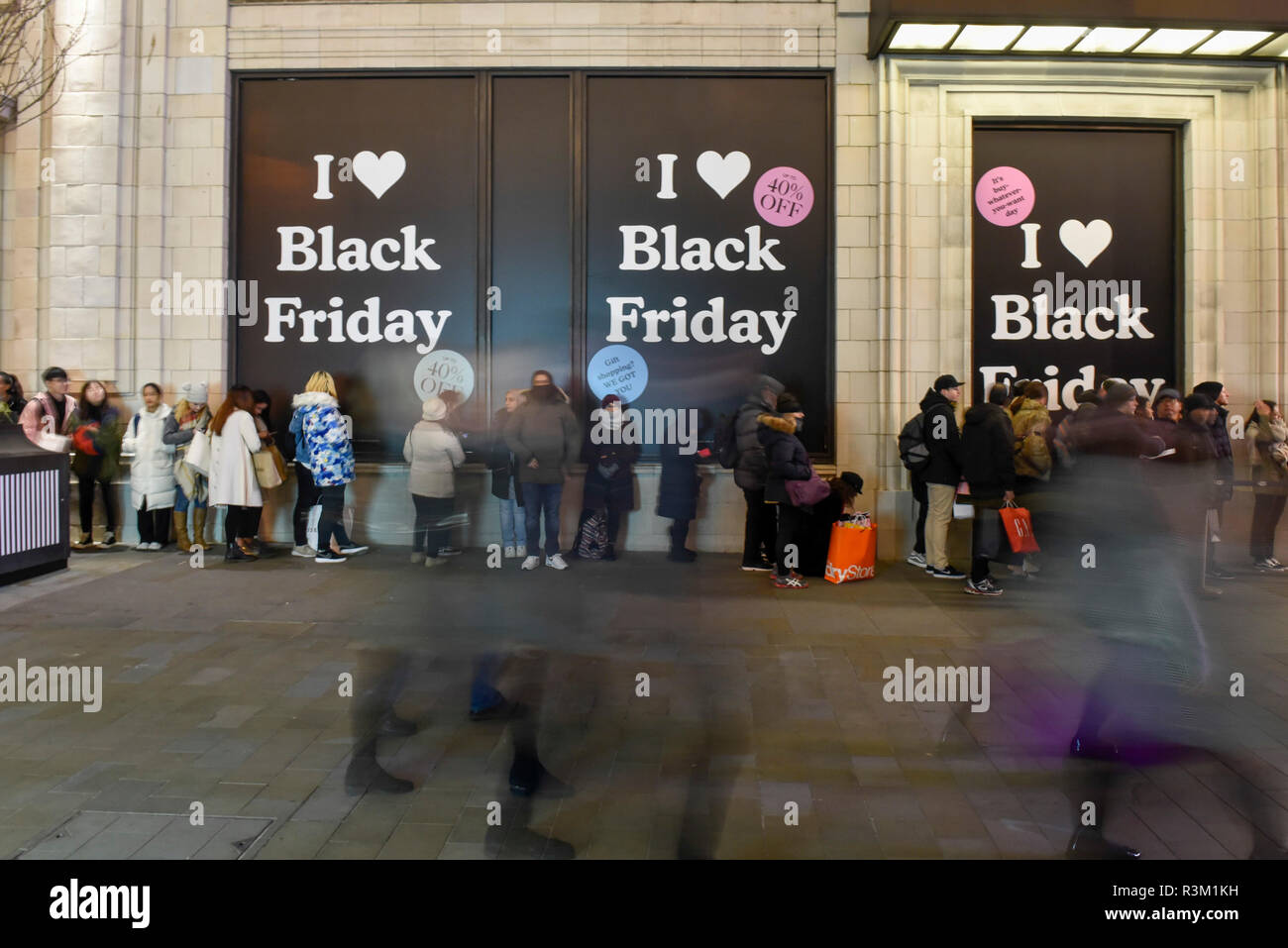London, UK. 23 November 2018. People queue into the night in front of  advertising signs outside the UGG store near Piccadilly Circus on Black  Friday. Traditional retailers face increasing challenges to attract