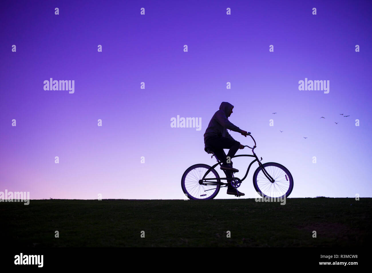 Silhouette of man in hoodie riding a beach cruiser bicycle against a glowing purple sky Stock Photo