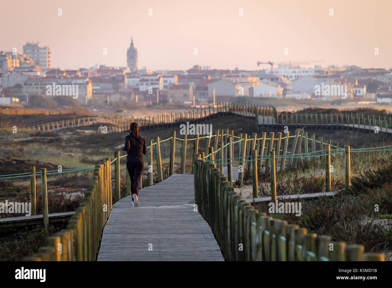 Young girl runs on boardwalk amidst vegetation towards distant city. Caucasian, dark hair, black track suit. Rear view. Copy Space. Clear day. Warm li Stock Photo