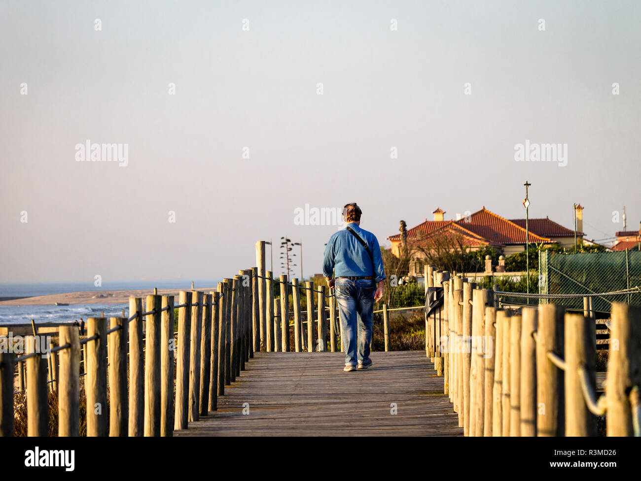 Middle-aged man walks on boardwalk near the ocean. Blue shirt, jeans. Rear view. Distant houses. Copy Space. Warm light. Stock Photo