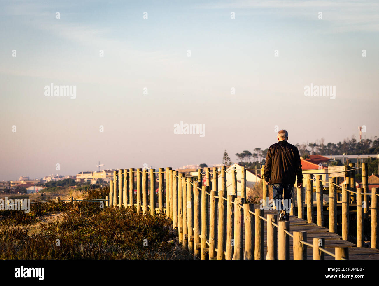 Senior man walks on boardwalk amidst vegetation. Brown coat, jeans. Rear view. Clear day. Copy Space. Stock Photo