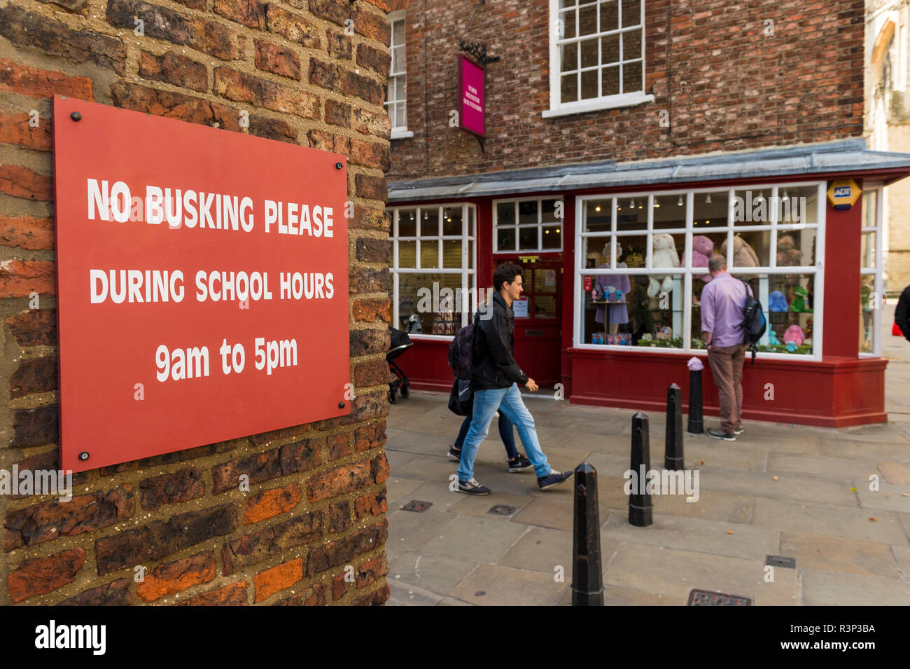People pass red sign attached to exterior brick wall, polite request saying 'no buskers please during school hours'  - York, Yorkshire, England, UK. Stock Photo