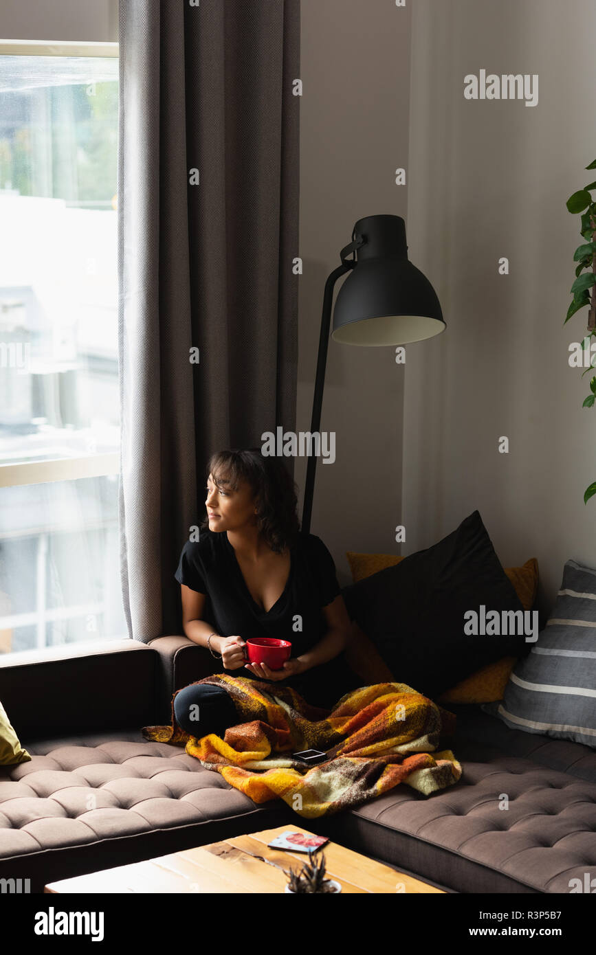 Woman looking through window while having coffee on sofa in living room Stock Photo