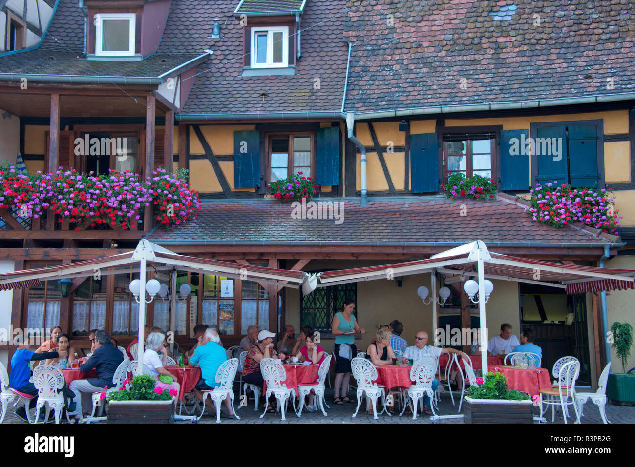 France, Alsace, Eguisheim. People fill an outdoor restaurant in front of a traditional building in Equisheim village. Stock Photo
