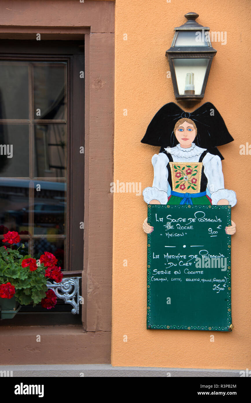 France, Alsace, Eguisheim. A picturesque sign with a traditionally dressed woman holding the chalkboard menu with offerings. Stock Photo