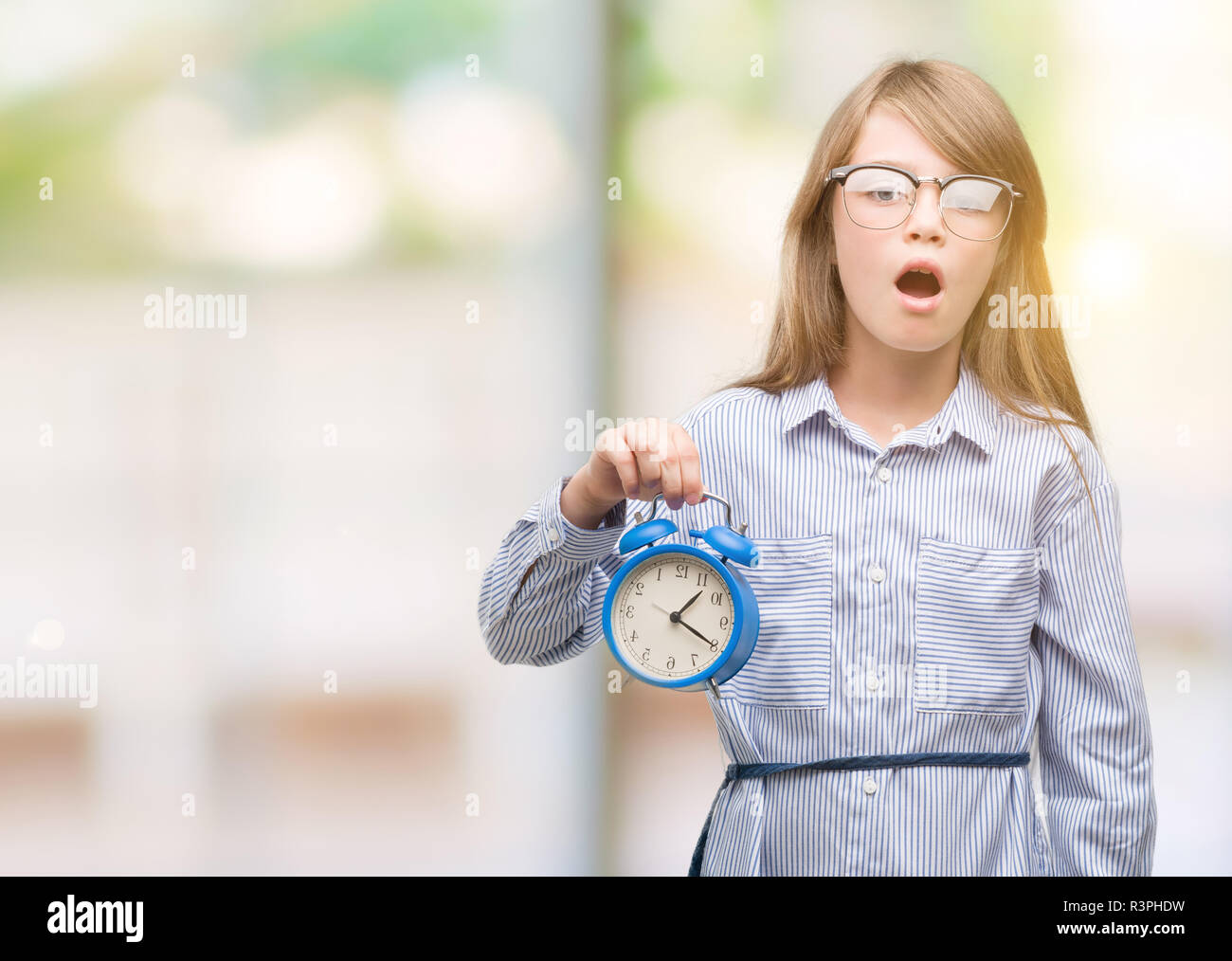 Young blonde child holding alarm clock scared in shock with a surprise