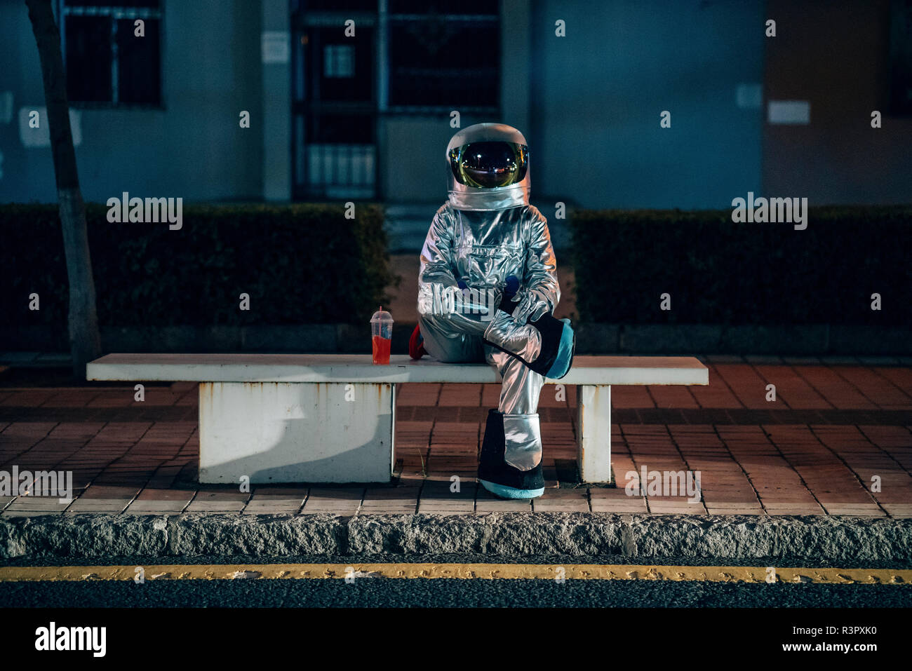 Spaceman sitting on bench at a bus stop at night with soft drink Stock Photo
