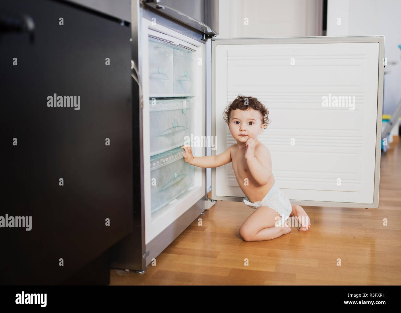 Portrait of baby boy wearing diaper crouching in front of refrigerator in the kitchen Stock Photo