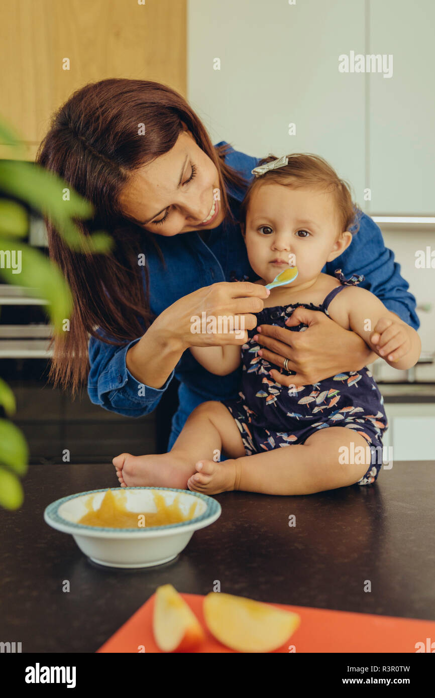 Mother helping baby daughter eating fruit pulp in kitchen Stock Photo