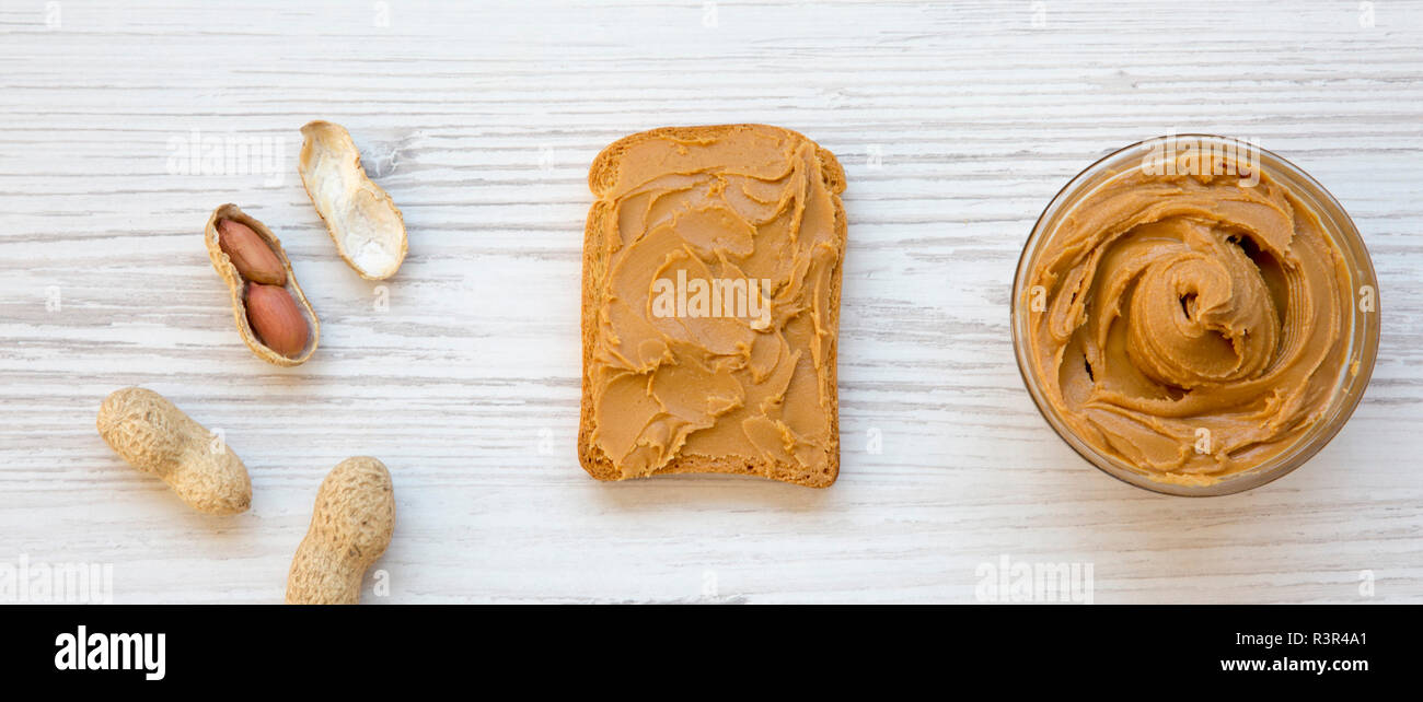 Toast with peanut butter, bowl of peanut butter and peanuts in shells on a white wooden surface, top view. Flat lay, overhead, from above. Stock Photo