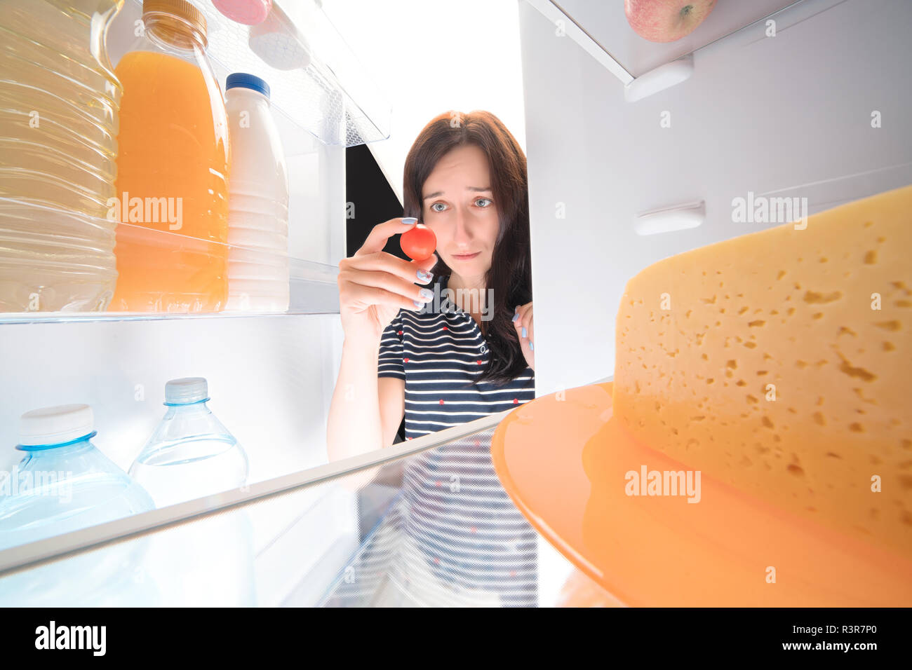 Sad girl looking into fridge and holding single tomatoes in her hand. Wide angle view from inside Stock Photo
