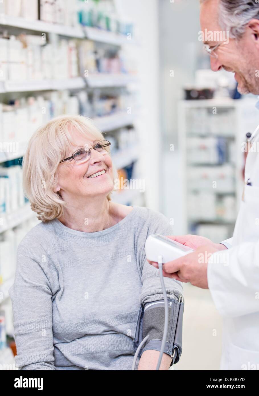 Male pharmacist checking smiling senior female patient's blood pressure in pharmacy. Stock Photo