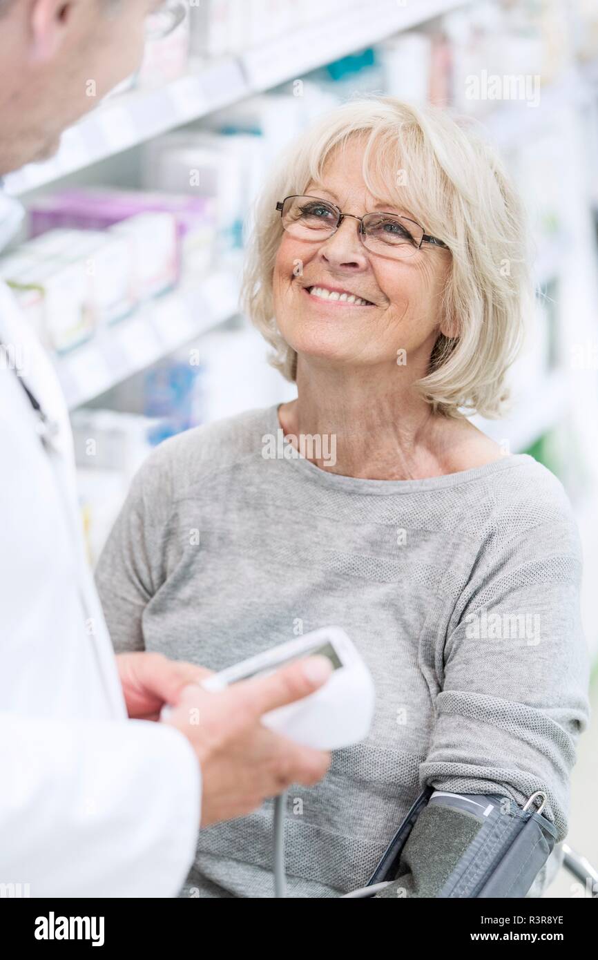 Male pharmacist checking smiling senior female patient's blood pressure in pharmacy. Stock Photo