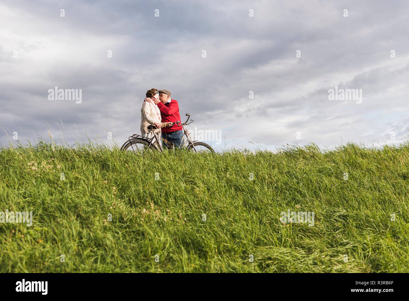 Senior couple with bicycles kissing in rural landscape under cloudy sky Stock Photo