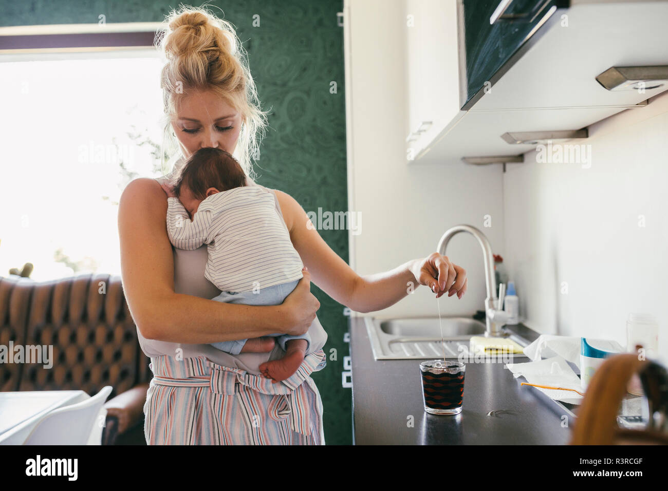Mother holding newborn baby in kitchen while making tea Stock Photo