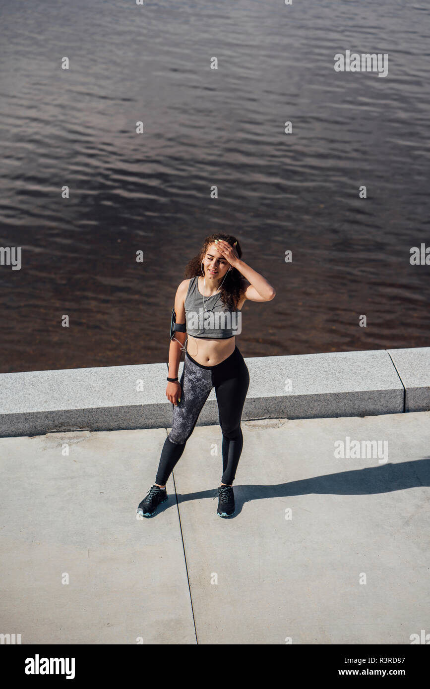 Exhausted young athletic woman wearing earbuds standing at the riverside Stock Photo