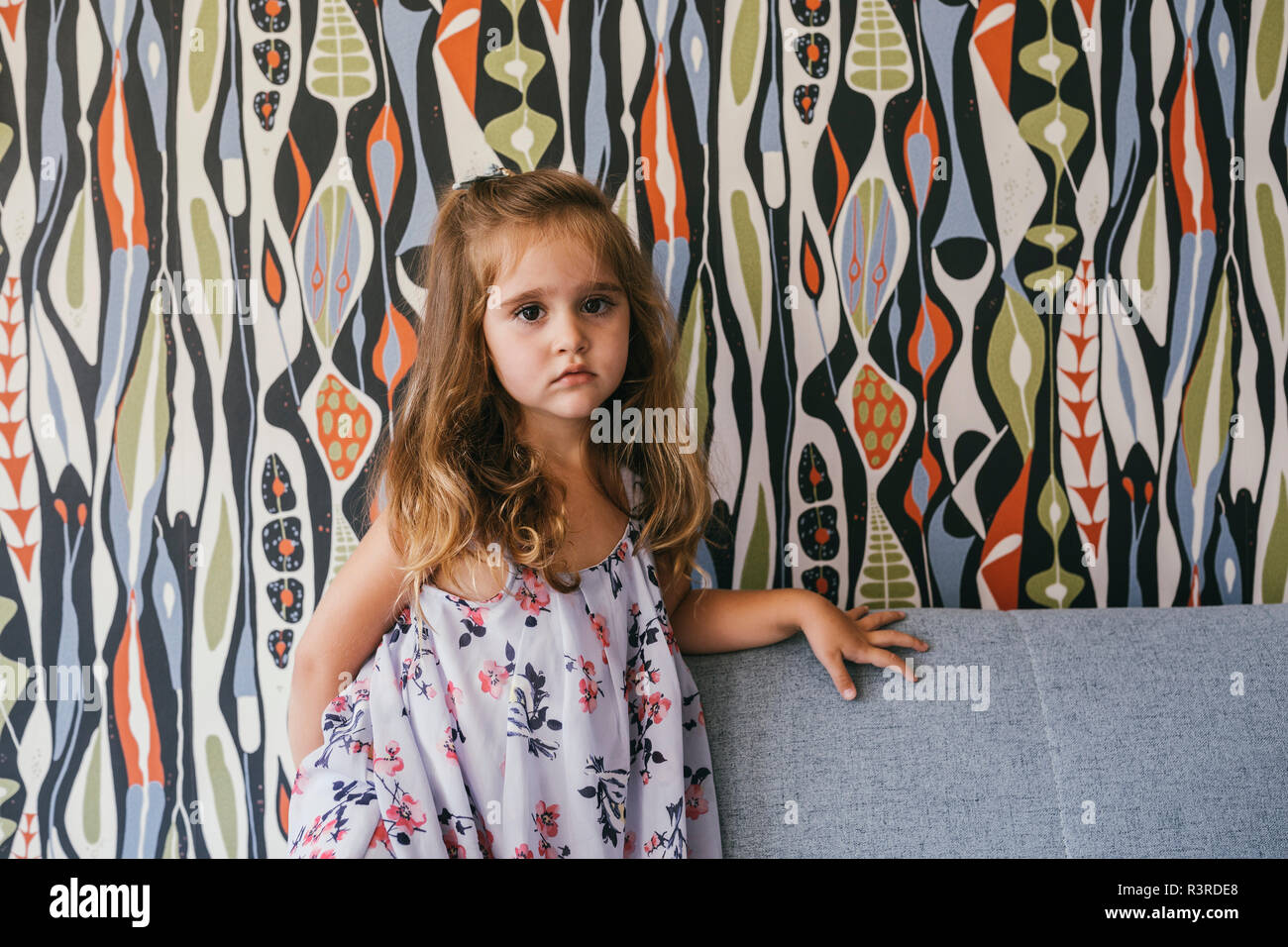 Portrait of serious little girl on couch in front of patterned wallpaper Stock Photo