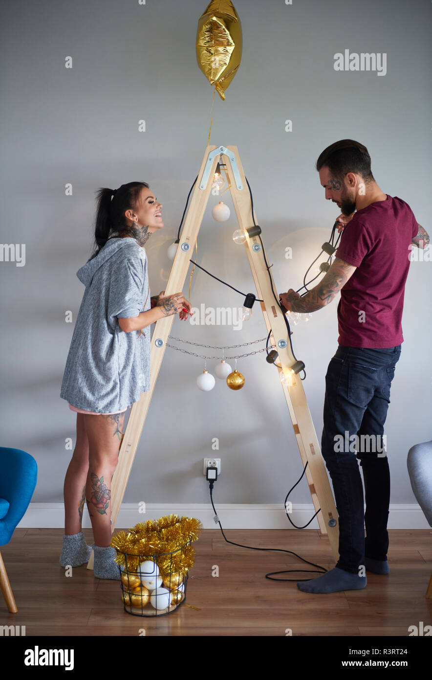 Modern couple decorating the home at Christmas time using ladder as Christmas tree Stock Photo
