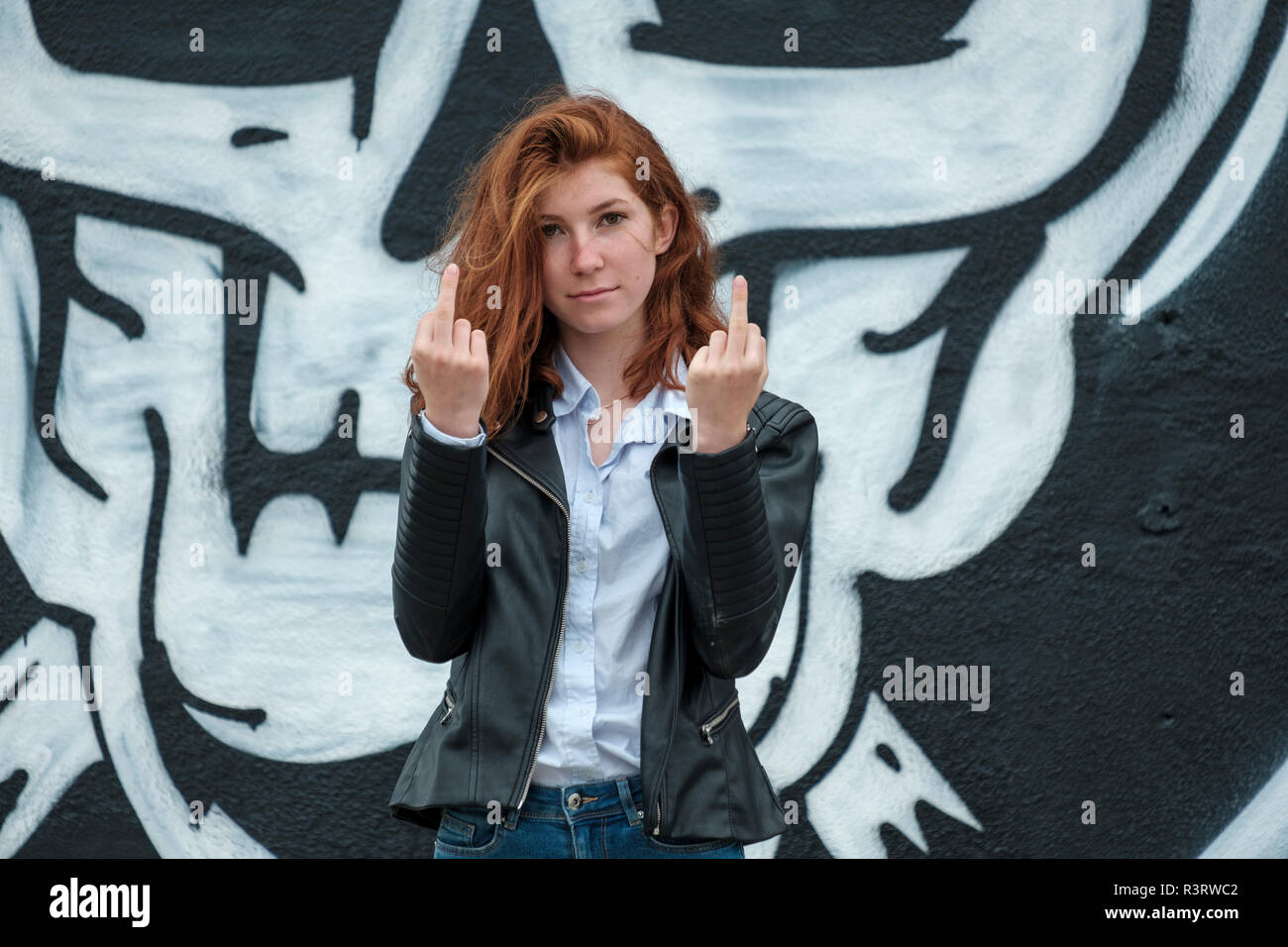 Italy, Finale Ligure, portrait of redheaded teenage girl showing the finger in front of wall with skull graffito Stock Photo