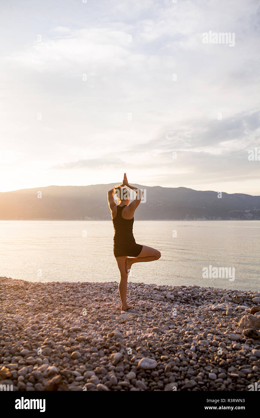 Young woman doing yoga on a stony beach at sunset, tree position Stock Photo