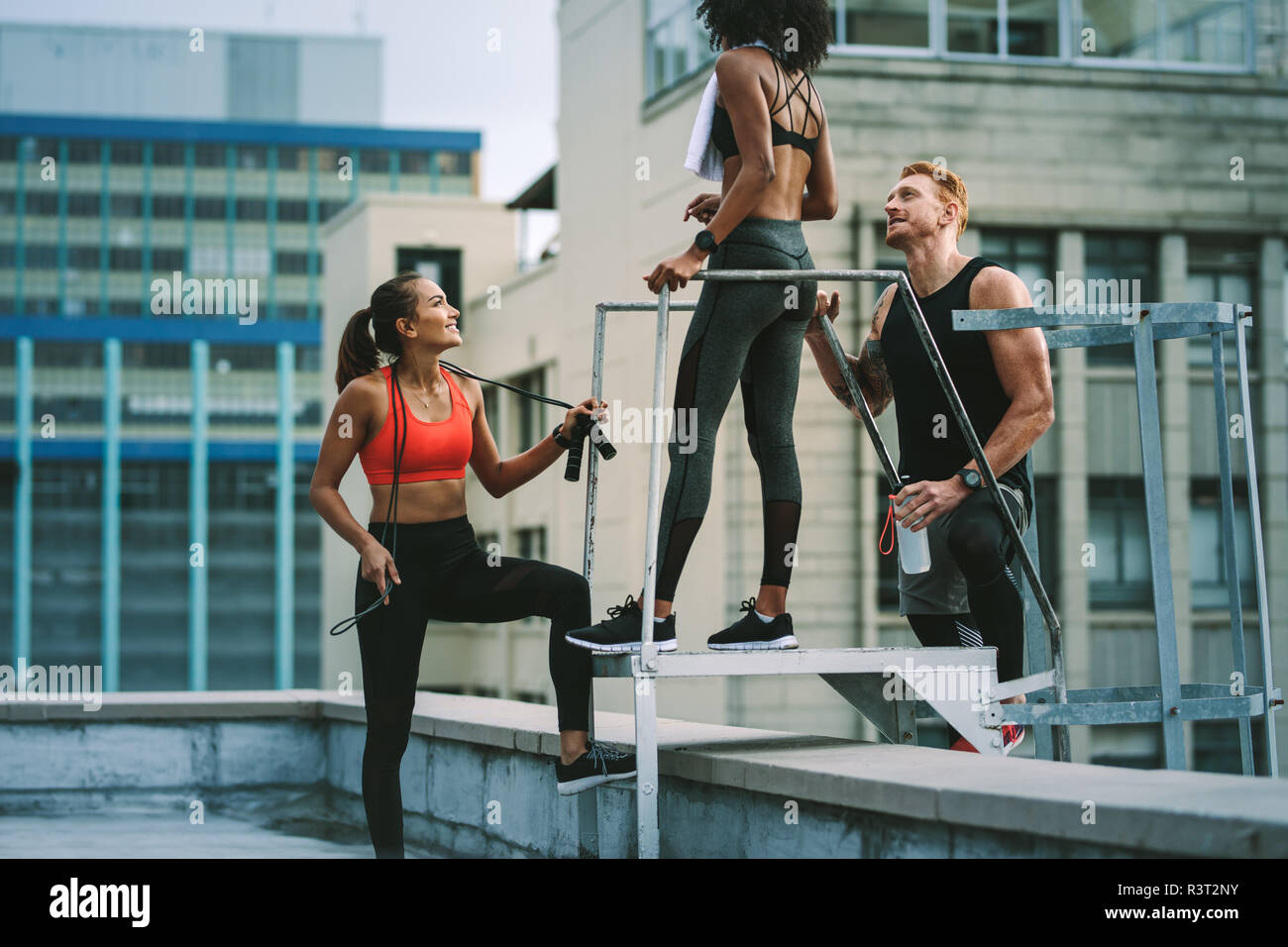 fitness woman standing on rooftop holding a skipping rope talking to her colleagues. fitness man standing with two fitness women on rooftop and talkin Stock Photo