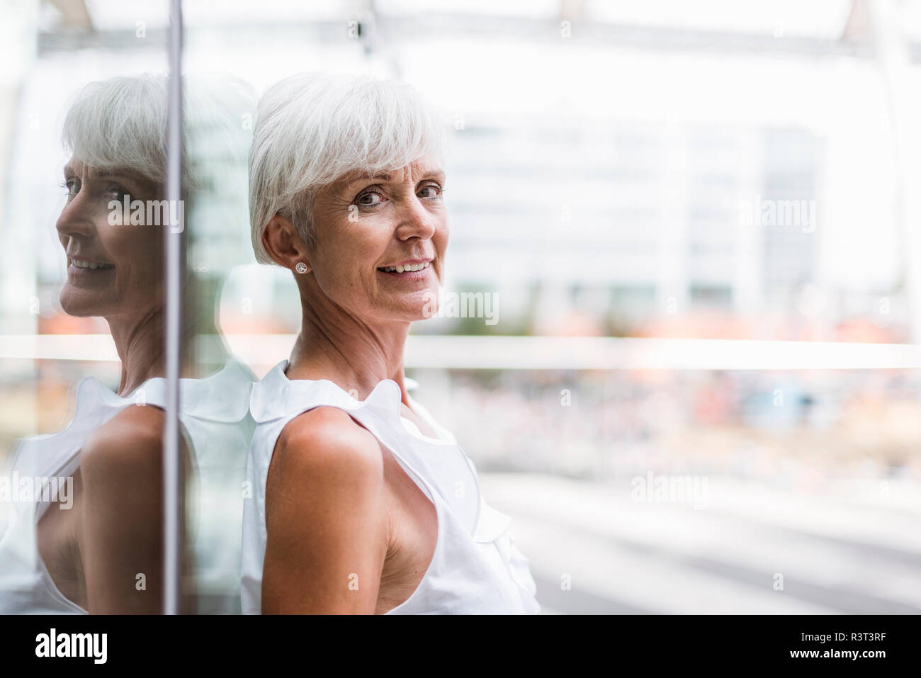 Portrait of smiling senior woman leaning against glass facade Stock Photo